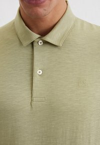 Oliwkowa koszulka polo wykonana z lekkiego materiału, z klasycznym kołnierzykiem, trzema guzikami oraz małym haftowanym logo na klatce piersiowej.