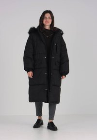 Abrigo puffer largo y negro con capucha y ribete de piel, cuenta con cierres de botones a presión, dos bolsillos frontales y un ajuste holgado.