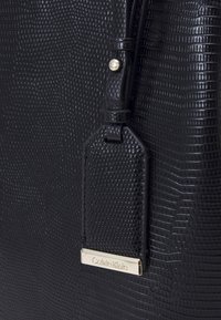 Sort tekstureret håndtaske med et reptil-lignende mønster, der har en guldfarvet logomærke og en læderrem, der fremviser elegante designdetaljer.