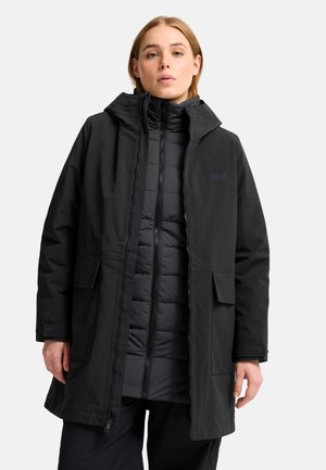 Donna che indossa un lungo cappotto invernale nero con cappuccio sovrapposto a una giacca imbottita nera, in piedi davanti a uno sfondo chiaro e uniforme.