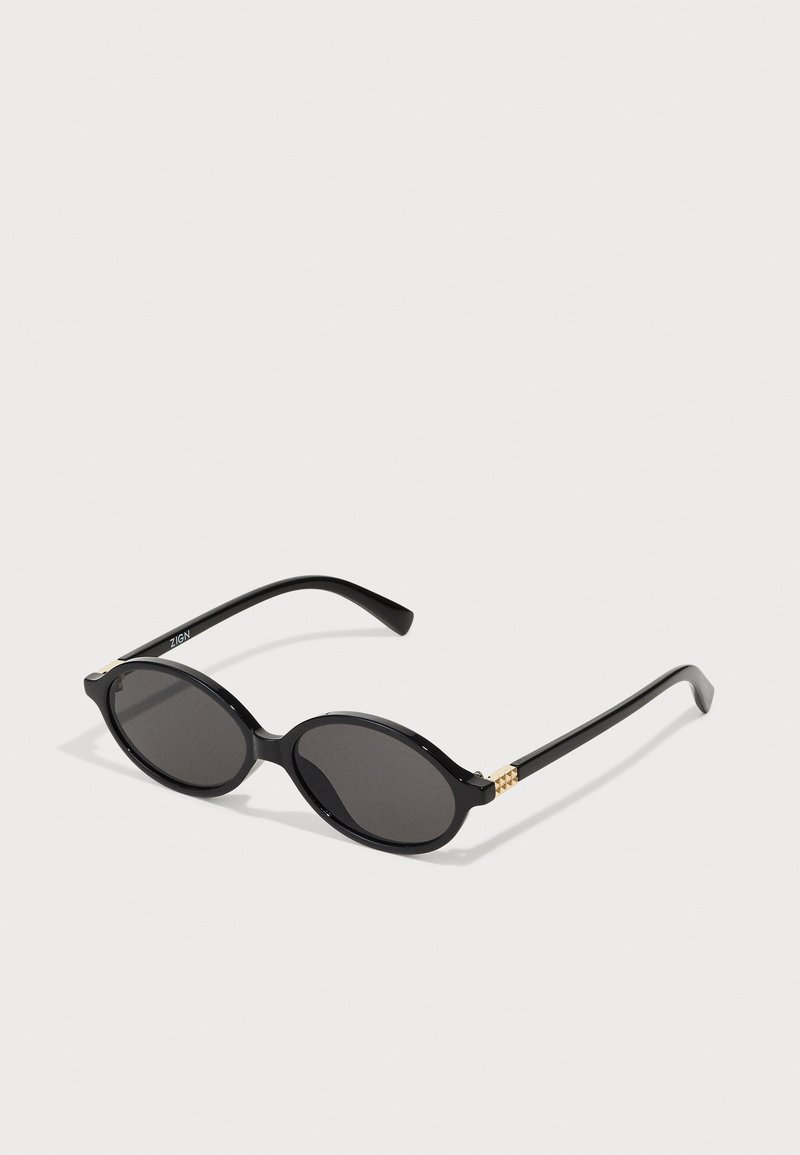 Óculos de sol ovais pretos com lentes escuras, armação de plástico brilhante e detalhes em dourado nas hastes.