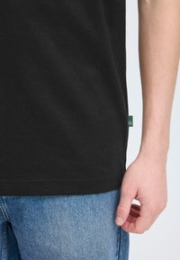 Tricou din bumbac negru cu o textură netedă, având un mic eticheta verde la tiv. Asortat cu blugi din denim de culoare albastră deschisă.