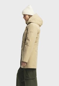 Giacca isolante beige con cappuccio, dotata di zip frontale e cordino. Abbinata a pantaloni cargo verde scuro. Berretto lavorato a maglia bianco.