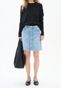 Kvinna iförd en svart långärmad topp och en ljusblå denimkjol med knäppning fram, som håller en stor svart handväska och bär svarta flats.