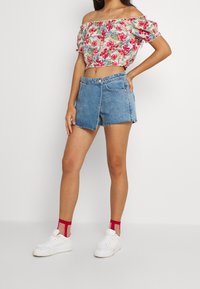 Donna che indossa un top corto floreale con spalle scoperte, pantaloncini di jeans asimmetrici, calze trasparenti rosse e sneakers platform bianche, in piedi con la mano sui fianchi.