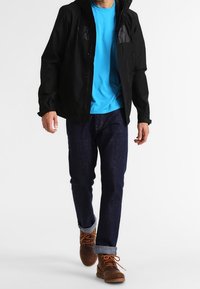 Schwarze Reißverschlussjacke mit Kapuze, blaues T-Shirt, dunkle Denim-Jeans und braune Schnürstiefel; mit Logos auf der Jacke und hochgekrempelten Jeansbündchen.