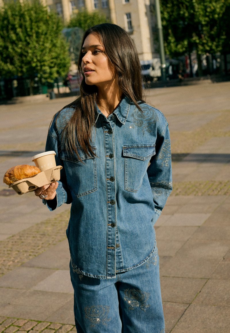 Jeune femme en veste et pantalon en jean tenant une tasse de café et un porte-croissants, debout à l'extérieur sur une place urbaine ensoleillée.