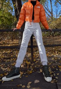 Chaqueta acolchada naranja, blusa negra, vaqueros blancos de cintura alta y zapatillas deportivas altas verdes con suelas blancas, contra un fondo otoñal con hojas.