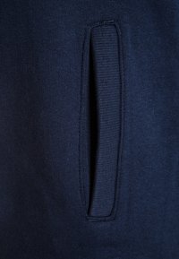 Primer plano de una tela azul marino con un bolsillo vertical y un ribete acanalado en un tejido de punto.