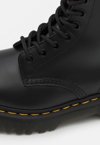 Stivale in pelle nera con punta tonda, suola in gomma spessa, lacci neri, cuciture gialle e occhielli. Presenta una texture liscia e un design robusto.
