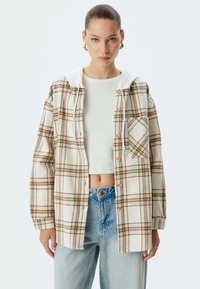 Kariertes Hemd in Creme und Braun mit einer weißen Kapuze, ausgestattet mit einer Knopfleiste und einer Brusttasche. Texturiertes Material mit lockerer Passform.