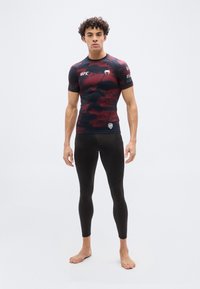 Νεαρός άνδρας, ξυπόλυτος, φορώντας στενό μαύρο και κόκκινο συμπιεστικό μπλουζάκι UFC και μαύρο κολάν, με φόντο απλό.