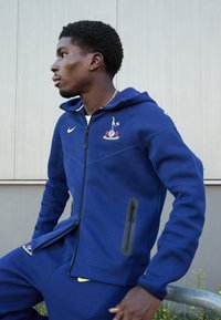 Marineblå fleece jubilumsgenser med frontlynlås, sidelommer og broderet logo. Medfølgende bukser med Nike-detalje. Flad tekstur, afslappet pasform.