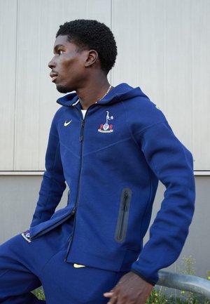 Navy blue fleece hoodie with a front zipper, side pocket, and embroidered logo. Accompanying pants with a Nike accent. Flat texture, casual fit.