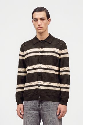 Jeune homme aux cheveux foncés coiffés en arrière, portant un cardigan en maille noir avec des rayures horizontales crème et un jean gris délavé.