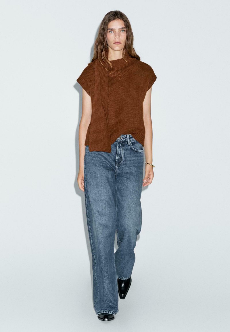Top in maglia marrone con scollo drappeggiato, abbinato a jeans a gamba larga blu chiaro e stivali con tacco neri. Tessuto semplice e strutturato in una vestibilità comoda.