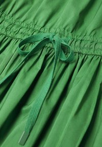 Zielony materiał sukienki zebrany w fałdy i z regulowanym sznurkiem w pasie. Materiał wydaje się gładki z delikatnym połyskiem.