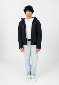 Zwarte zip-up jack met capuchon, voorzien van voorkruiszakken en een soepele textuur, gedragen over een witte t-shirt en lichtblauwe jeans.