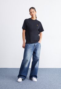 Sort bomulds t-shirt med korte ærmer og et lille gult logo, parret med løse blå jeans og hvide sneakers på en struktureret overflade.