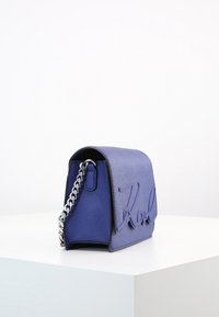 Modrá kožená kabelka přes rameno se stříbrným řetízkovým popruhem a vyraženým nápisem na předním klapce, vystavená na bílé ploše.