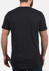 Schwarzes Baumwoll-T-Shirt mit kurzen Ärmeln, das auf dem Rücken ein wiederholtes weißes Diamantenmuster aufweist. Rundhalsausschnitt-Design mit lockerer Passform.
