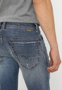 Blå denimjeans med en figurnära design, med slitage på fickorna och en märkeslogg. Bakvyn visar detaljerad sömnad och textur.