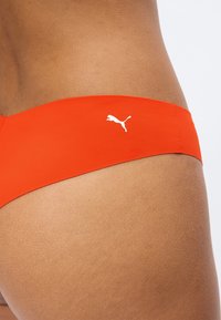 Κοντινή λήψη του ισχίου ενός ατόμου που φοράει πορτοκαλί μαγιό μπικίνι Puma με λευκό λογότυπο Puma στο πλάι.