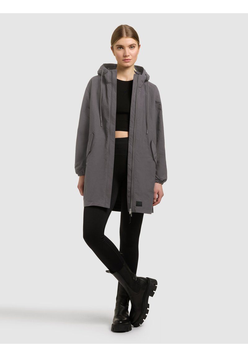 Jeune femme debout, jambes croisées, portant une veste à capuche grise, un crop top noir, des leggings noirs et des bottes épaisses noires.
