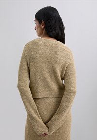 Woman with long dark hair stands facing away, wearing a beige textured knit sweater and matching skirt, hands clasped behind her back.
