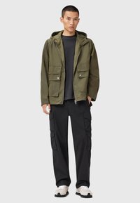 Veste à capuche vert olive avec une fermeture éclair, dotée de deux poches poitrine ; associée à un pantalon cargo noir et des baskets beiges texturées.