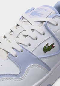 Άσπρο και γαλάζιο αθλητικό παπούτσι με λευκά κορδόνια και πράσινο κεντητό λογότυπο κροκόδειλου στο πλάι κοντά στη φτέρνα.