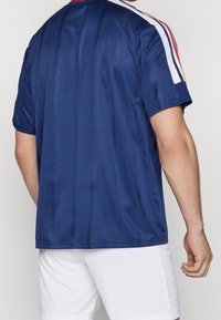 Man som bär en marinblå kortärmad sportskjorta med röda och vita ränder på ärmarna och vita träningsshorts, vänd bort.