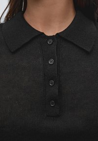 Mustapaitainen neulepaitapolo, jossa on kaulus ja kuusi mustaa nappia napituksessa, sekä teksturoitu kangas.