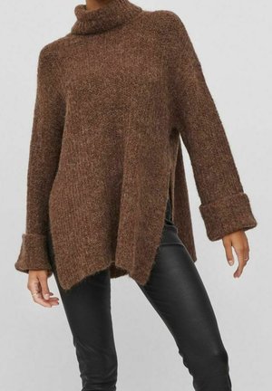 Femme portant un pull à col roulé marron oversize avec des fentes latérales et un pantalon noir ajusté sur un fond uni.