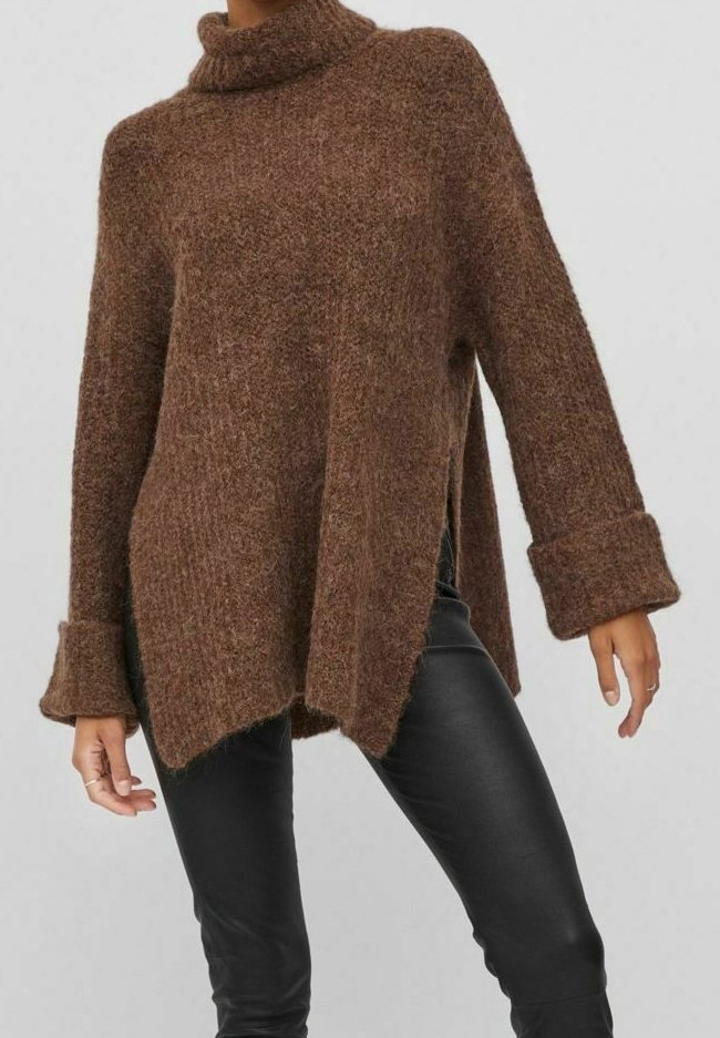 Femme portant un pull à col roulé marron oversize avec des fentes latérales et un pantalon noir ajusté sur un fond uni.