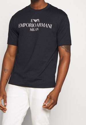 Homme portant un t-shirt noir avec le logo Emporio Armani Milan et un pantalon blanc, avec des tatouages visibles sur les deux avant-bras.