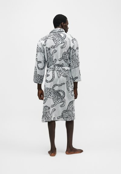 Άνδρας που φοράει λευκή ρόμπα μήκους μέχρι το γόνατο με μαύρα λεοπάρ σχέδια, όρθιος ξυπόλυτος με την πλάτη προς έναν απλό φόντο.