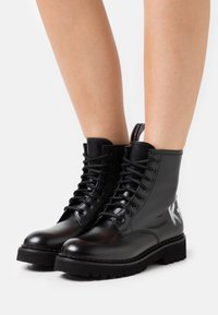Botas de combate de cuero negro con un acabado suave, con cordones en la parte delantera, suelas resistentes y un detalle en forma de "K" estilo graffiti en el costado.