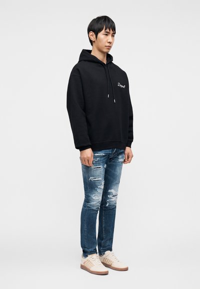 Μαύρο φούτερ με κουκούλα και τσέπη καγκουρό μπροστά, συνδυασμένο με φθαρμένο μπλε τζιν και μπεζ αθλητικά παπούτσια με καφέ λεπτομέρειες.