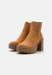 Smeđi suede ankle čizme s visokim blok petama i debelim, rebrastim potplatom. Imaju elastične bočne panele i prošivene akcentne detalje oko potplata.