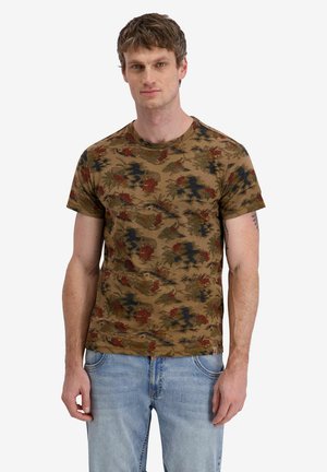 Man wearing a brown floral camouflage patterned t-shirt and light blue jeans standing against a plain background.