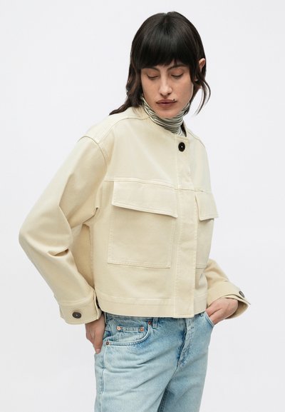 Γυναίκα με σκούρα μαλλιά που φοράει κρεμ κοντό μπουφάν πάνω από ριγέ ζιβάγκο και ανοιχτό μπλε τζιν, με τα χέρια στις τσέπες, κοιτάζοντας προς τα κάτω.