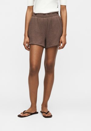 Pantaloncini marroni con texture e vita elasticizzata indossati con top bianco e infradito marroni su persona in piedi su sfondo semplice.