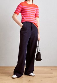 Punainen ja vaaleanpunainen raidallinen neulepaita lyhyillä hihoilla, yhdistettynä tummansinisiin leveälahkeisiin housuihin. Mallilla on pieni musta laukku käsissään ja päällään valkoiset lenkkarit.