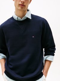Junger Mann trägt einen dunkelblauen Pullover über einem hellblauen Hemd mit hochgekrempelten Ärmeln, Hände in den Taschen, vor weißem Hintergrund.