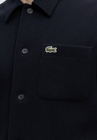 Veste en laine bleu marine foncé avec une poche avant ornée d'un petit logo crocodile vert et trois boutons noirs sur le devant.