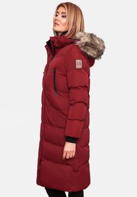 Red padded parka with fur-lined hood, zippered pockets, and contrasting black accents. Features a logo patch on the arm and a long silhouette.