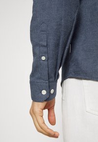 Niebieska koszula z długim rękawem w kratę, z białymi guzikami i detalami przy mankietach. Tkanina wydaje się być teksturowana, co tworzy wzorzysty efekt wizualny.