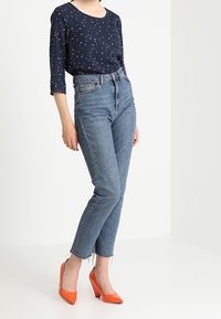 Blusa blu navy con pois bianchi e rosa, jeans in denim blu a vita alta e scarpe con tacco arancioni a punta.