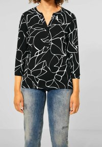 Svart topp med vita abstrakta linjemönster, trekvartsärmar och notch-krage. Buren med slitna blå jeans.